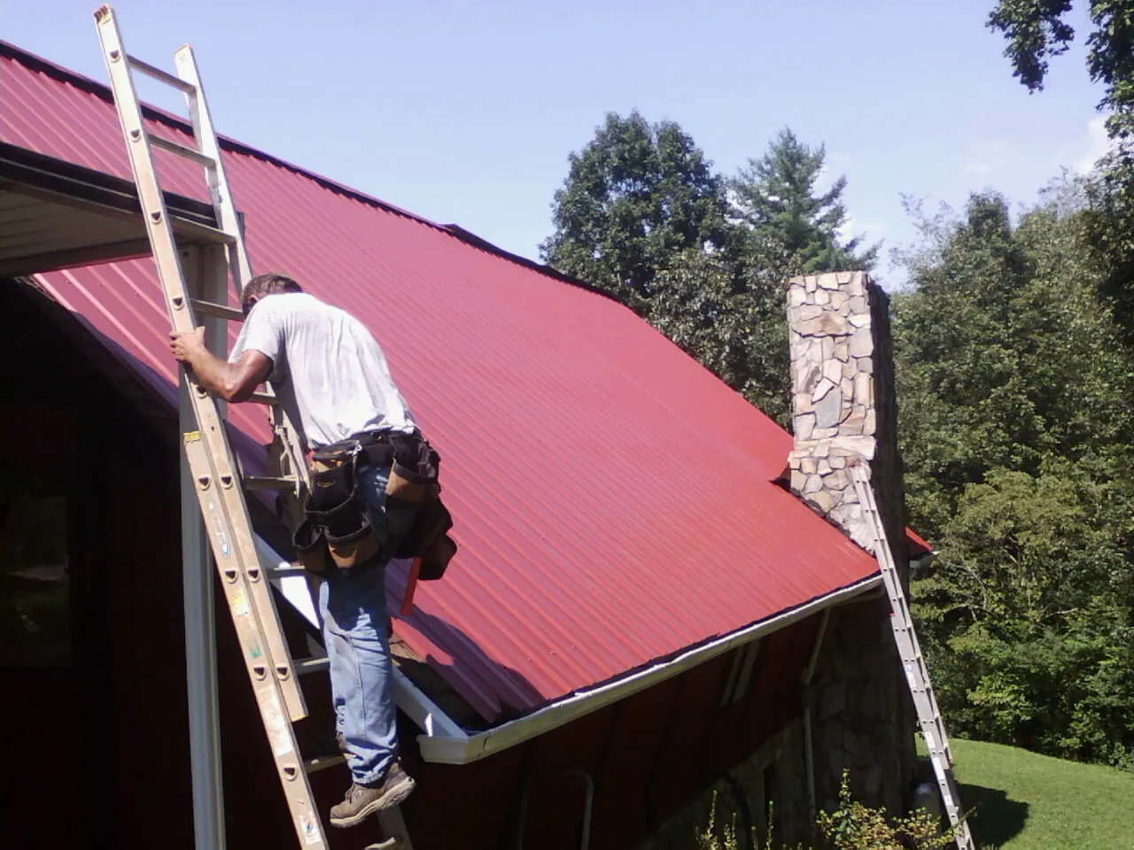 Roofer on a ladder beside a red metal roof and stone chimney — Soffit & Fascia Repair by Roofing Brothers in Beardsley
